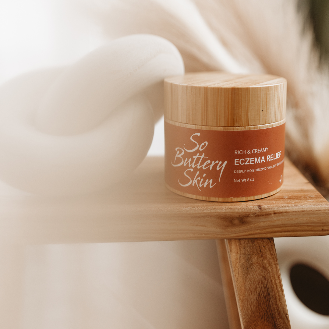 So Buttery Skin rich and creamy eczema relief body butter with a bamboo lid, displayed on a wooden stool alongside a modern white knot pillow
