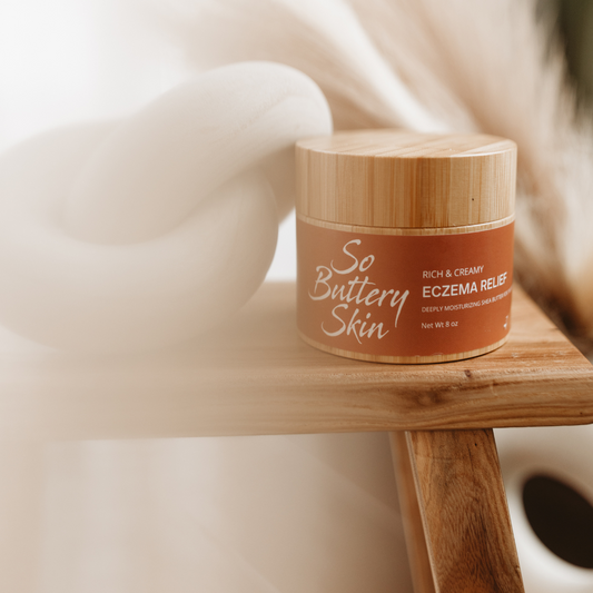 So Buttery Skin rich and creamy eczema relief body butter with a bamboo lid, displayed on a wooden stool alongside a modern white knot pillow